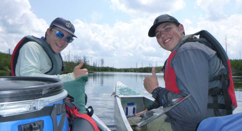 Two people wearing life jackets sit inside canoes and give the camera a thumbs up